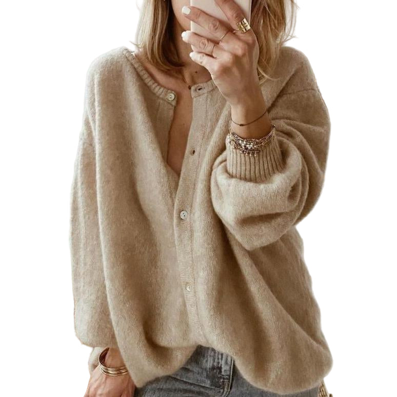 Une femme prend une photo dans un miroir, vêtue d'un cardigan marron oversize à boutons, en tissu doux et duveteux, associé à un jean décontracté et à plusieurs bracelets en or, dans une chambre lumineuse à la décoration moderne.
